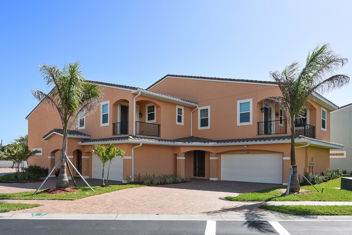 A two-story townhome features a warm coral exterior complemented by a tiled roof. Two palm trees provide shade in the front yard, while a spacious driveway leads to two garage doors. The well-maintained landscaping adds to the inviting curb appeal.