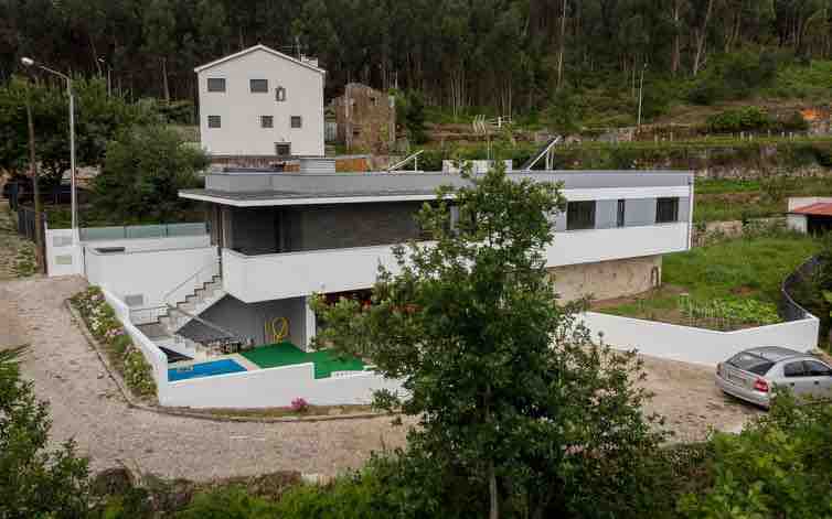 The modern building is set against a backdrop of greenery, featuring multiple levels and large windows. A stone pathway leads to the entrance, where a swimming pool and outdoor area are visible. Surrounding vegetation creates a natural, serene environment.