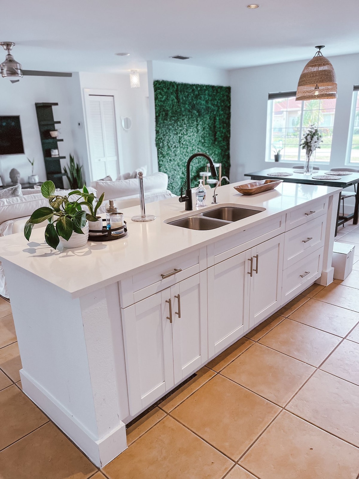 A modern kitchen island features a white countertop and spacious cabinetry. A stainless steel sink is positioned alongside a sleek faucet. In the background, greenery adorns the wall, while natural light streams through large windows, illuminating the inviting space.