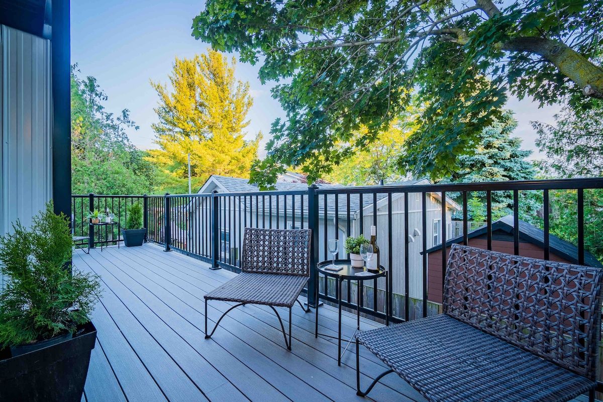 An outdoor deck space is presented with two woven chairs and a small round table. Lush trees provide a natural backdrop, with glimpses of neighboring houses in the distance. The deck's wooden flooring reflects a modern design, creating a serene area for relaxation.