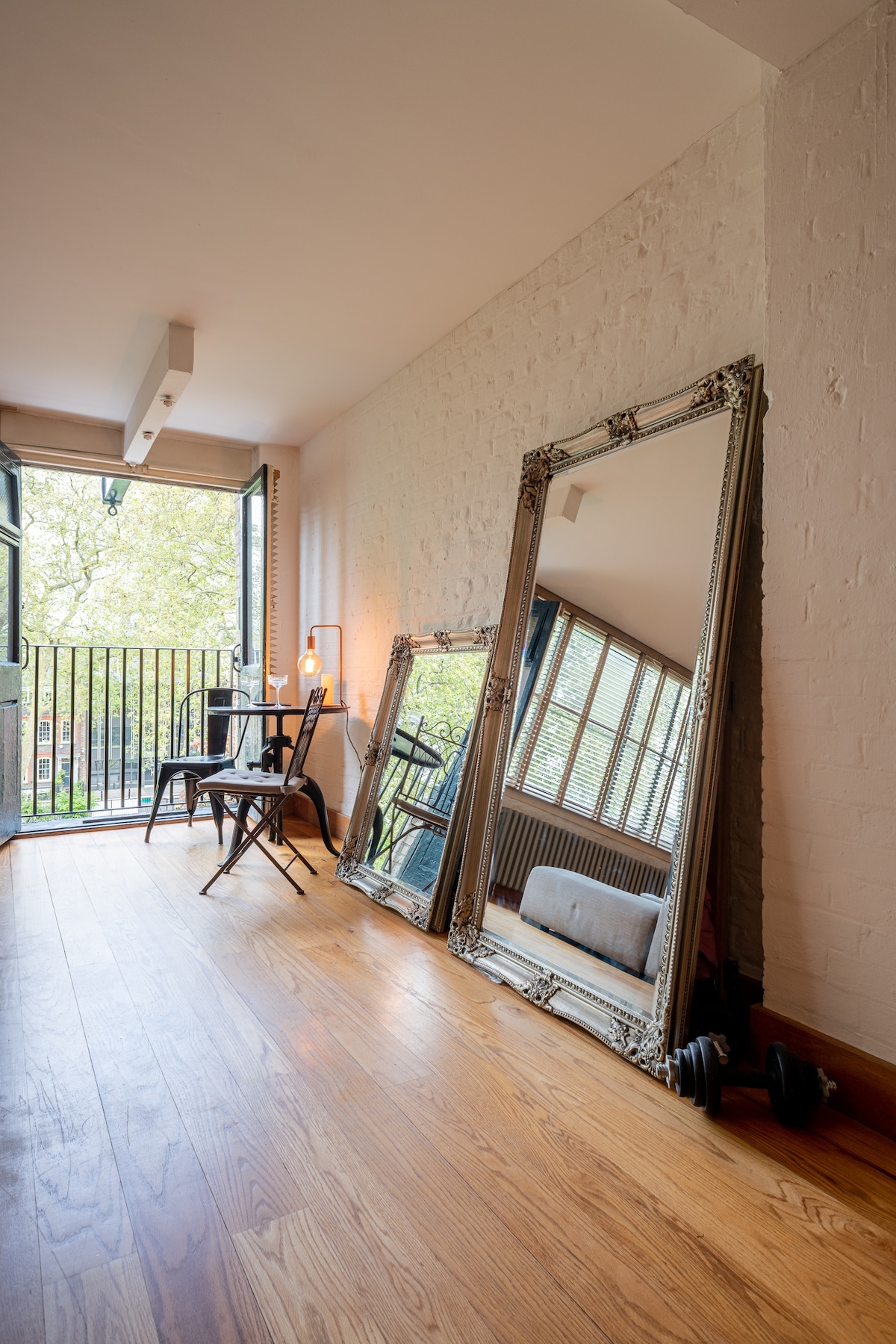 An elegant corner of the loft space features twin tall mirrors leaning against the wall, reflecting natural light from large windows. A small bistro table and chair are positioned nearby, with a lamp providing warm illumination. Wooden flooring adds warmth and texture to the space.