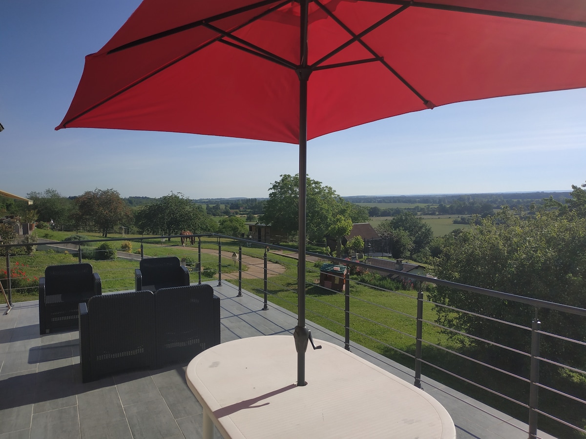 A spacious outdoor terrace is seen, featuring a red umbrella providing shade over a light-colored table. Comfortable seating is arranged nearby, with scenic views of the lush green countryside and distant horizon in the background.