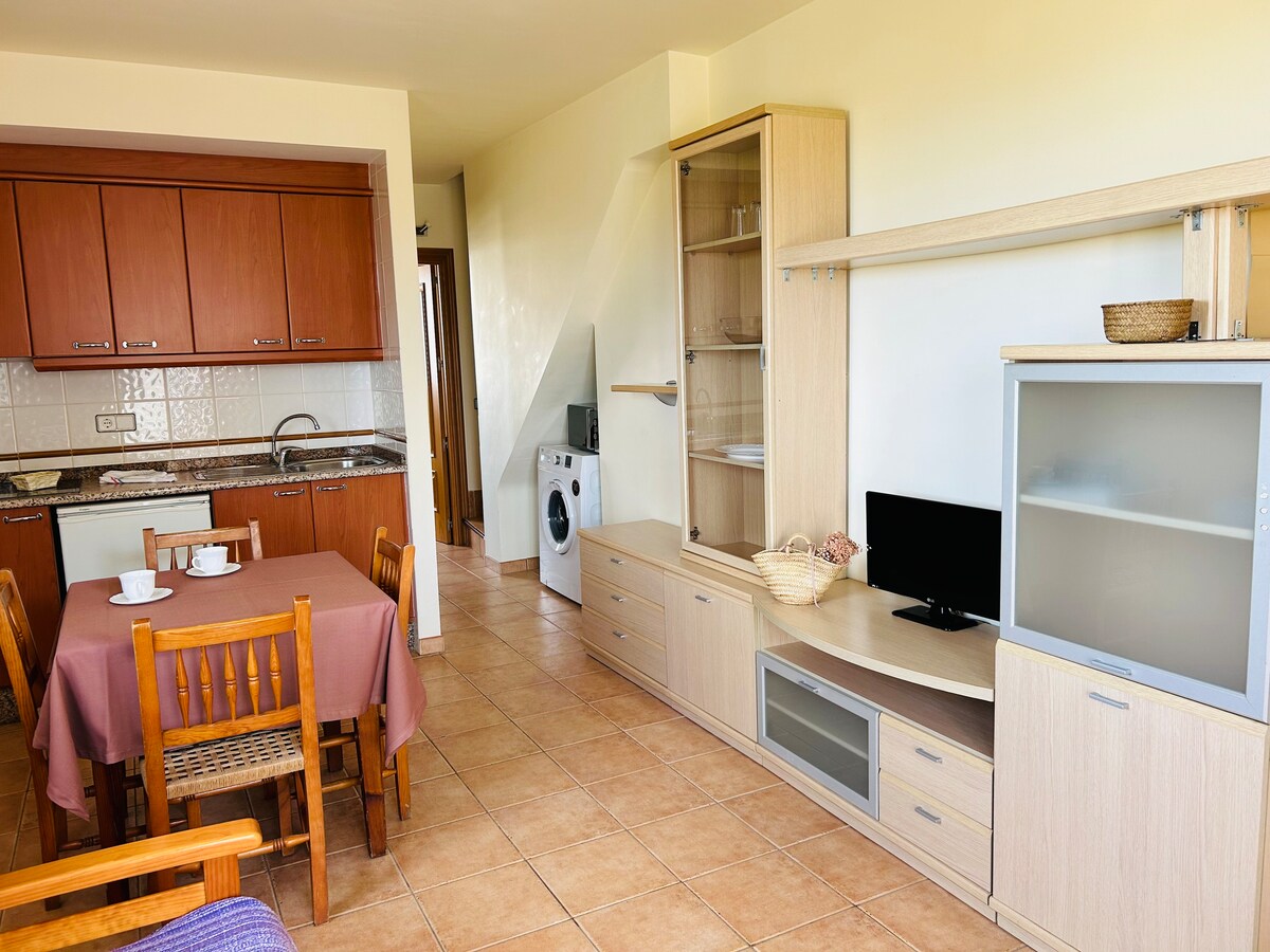 An open-plan living area features a wooden dining table with matching chairs and a kitchenette equipped with cabinetry. A small television sits on a light-hued entertainment unit, beside a wicker basket. The room is illuminated by natural light from a window.