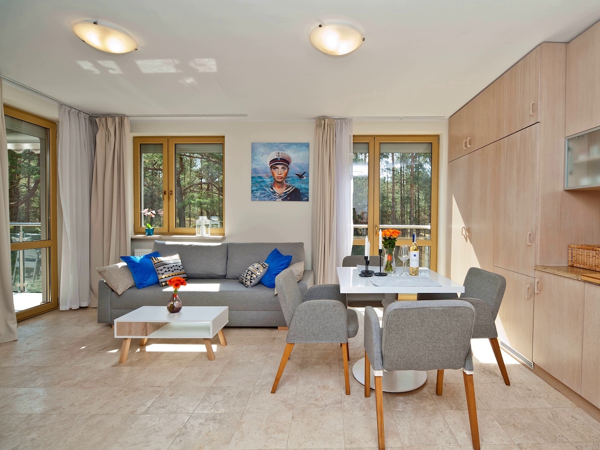 A light-filled living area features a comfortable gray sofa adorned with colorful cushions, complemented by a small coffee table. A dining table with four chairs occupies the space, surrounded by large windows that offer views of the outdoors. A floral arrangement adds a touch of color.