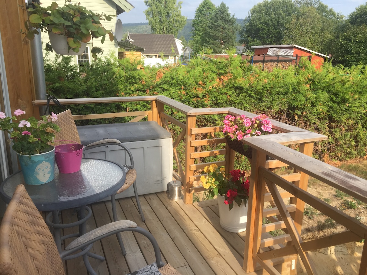 A wooden deck is shown, featuring a small round table with two chairs. Planters with colorful flowers are positioned on the table and at the railing, surrounded by greenery that offers a sense of privacy. A storage bench is visible along the side.
