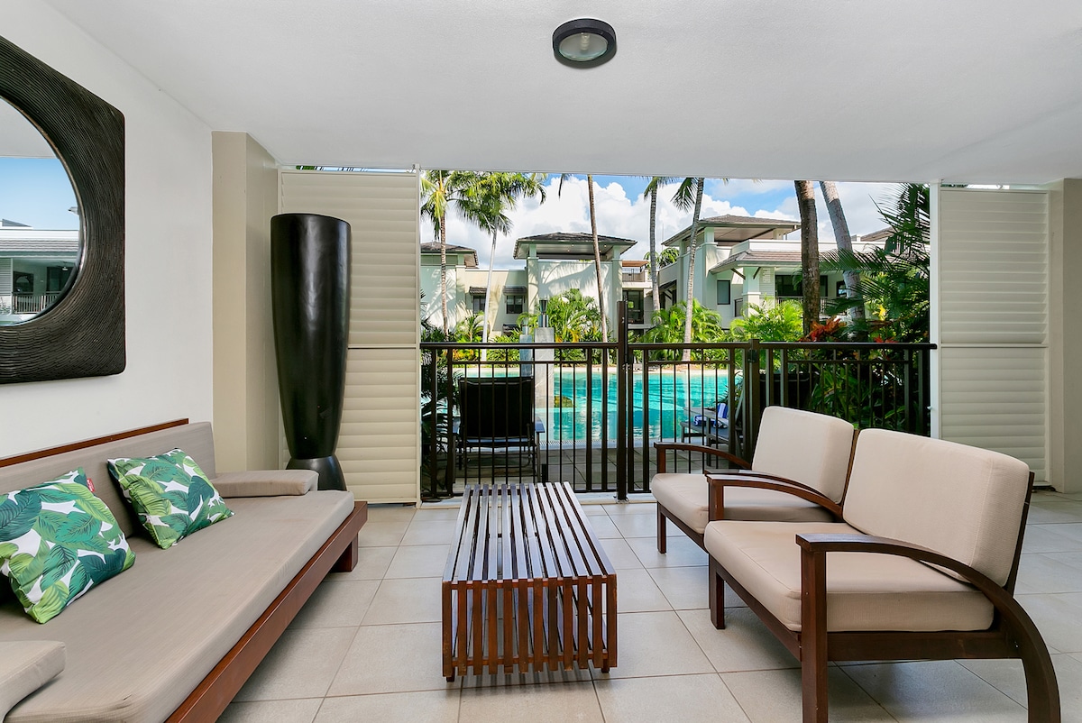 An inviting outdoor seating area features a wooden table flanked by two armchairs and a built-in sofa adorned with tropical cushions. Glass doors provide a view of the resort pool and lush surroundings, while partial walls allow for airflow and natural light.