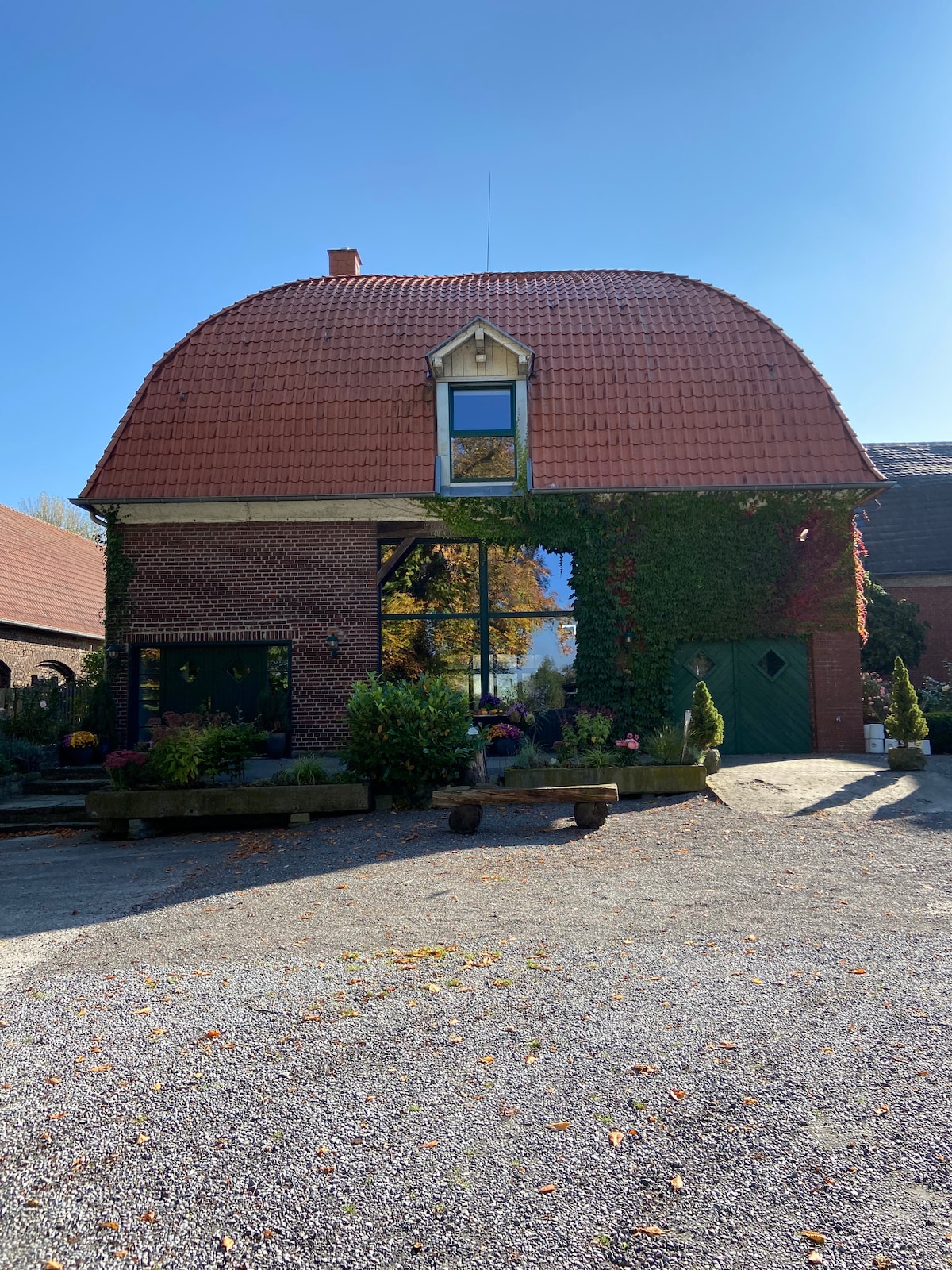 A charming brick building features a distinctive curved roof and large windows reflecting the surrounding greenery. The facade is adorned with climbing plants and colorful flowers, while well-maintained pathways lead to inviting entrances. Clear blue skies create a serene backdrop.