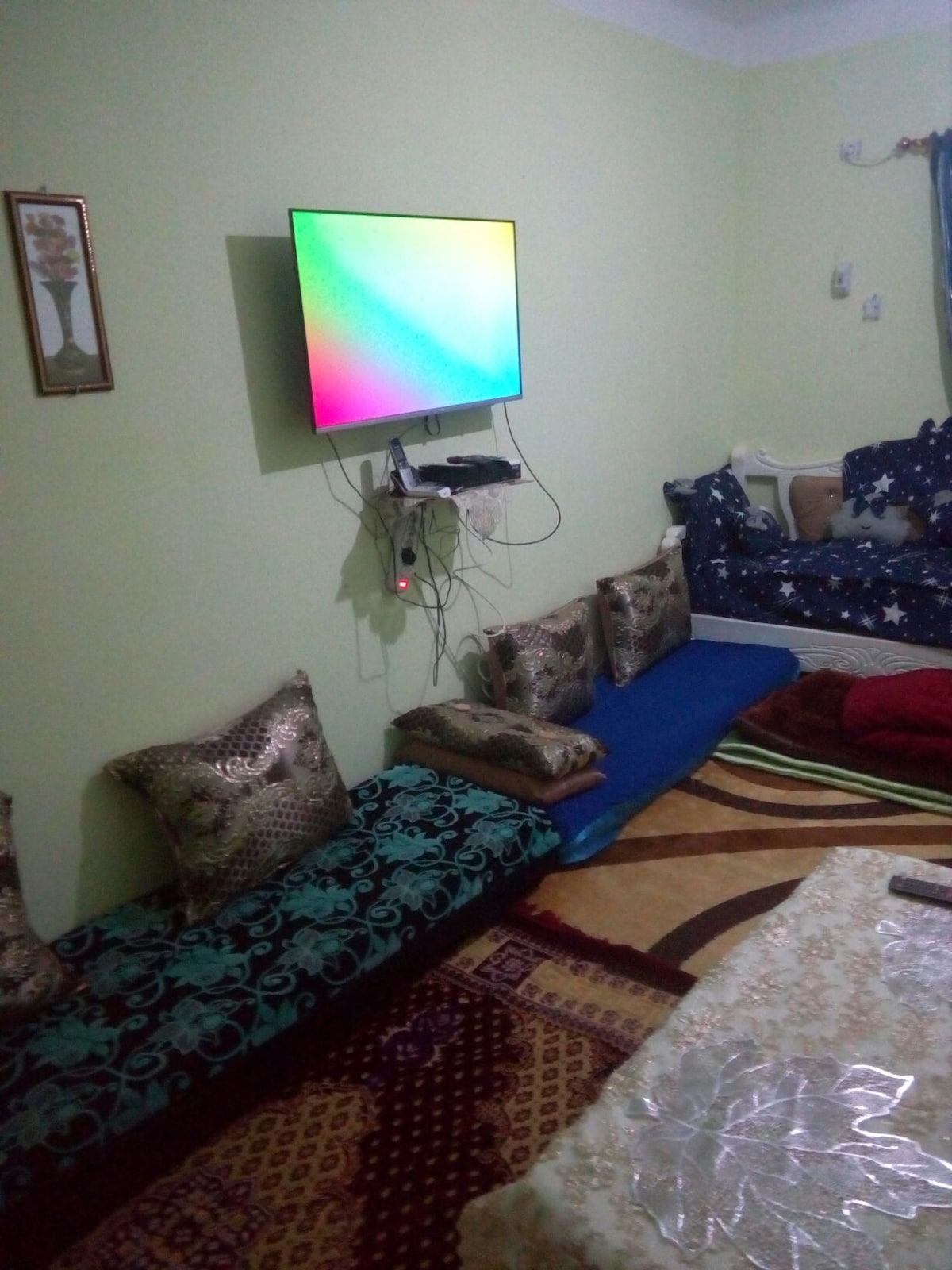 A living area is depicted with a mounted television on a light green wall. Low seating in various colors is arranged against the wall, adorned with decorative cushions. A patterned area rug covers the floor, and a glimpse of a bed is visible in the corner.