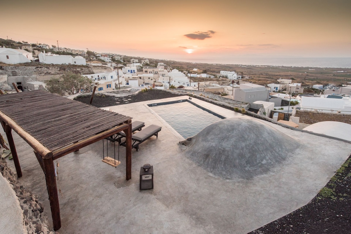 An outdoor space showcases a tranquil swimming pool surrounded by smooth, sunlit concrete surfaces. A wooden pergola and a sun lounger provide relaxation options. In the background, traditional white buildings cluster along the hillside, with the sun setting over the distant landscape.