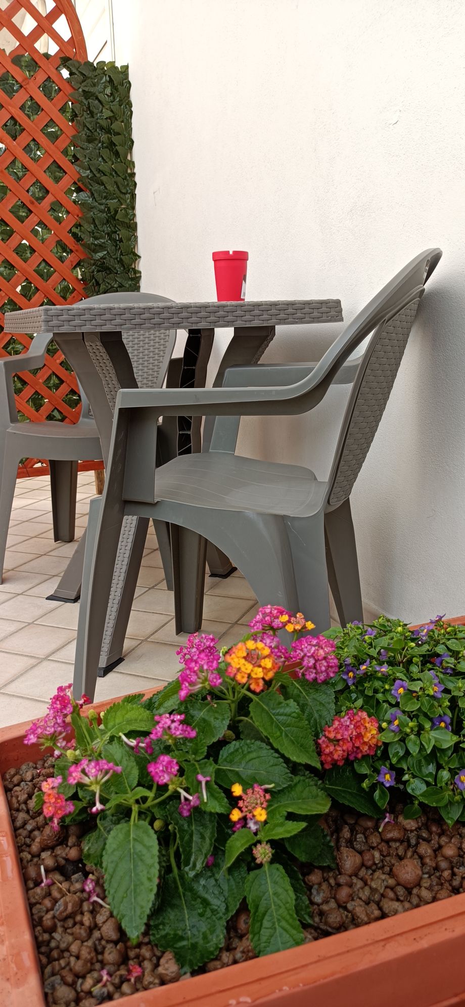 An outdoor seating area features a gray plastic table and chairs set against a white wall. A vibrant display of flowers in shades of pink, orange, and purple is planted in the foreground, adding a touch of color to the space.