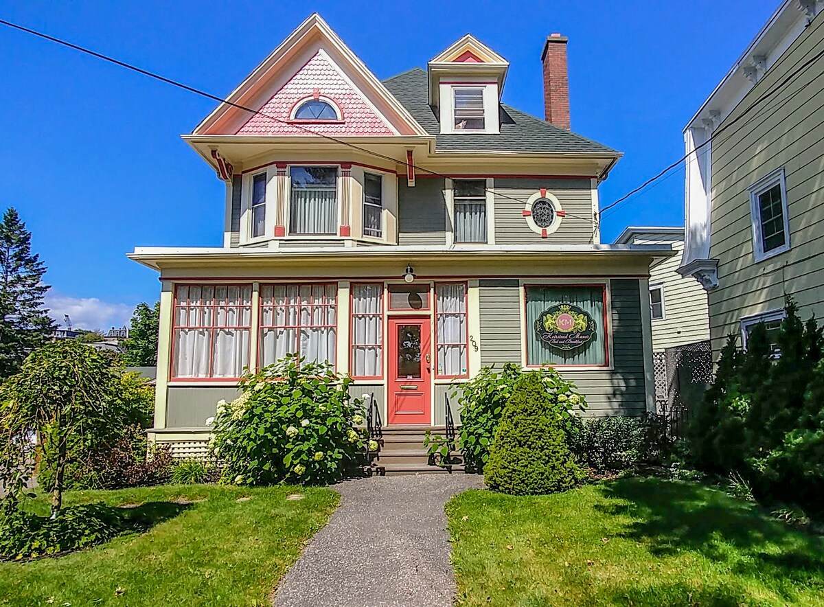 A charming boutique-style B&B is showcased with its distinctive architecture featuring a gabled roof and decorative trim. Lush greenery surrounds the pathway leading to a vibrant red door, which is framed by large windows that allow natural light to fill the welcoming entrance.