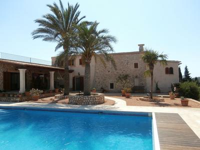 A stone building is set against a clear blue sky, surrounded by palm trees and terracotta pots. A swimming pool in the foreground reflects the sunlight, while a spacious terrace with shaded areas can be seen along the side of the property.