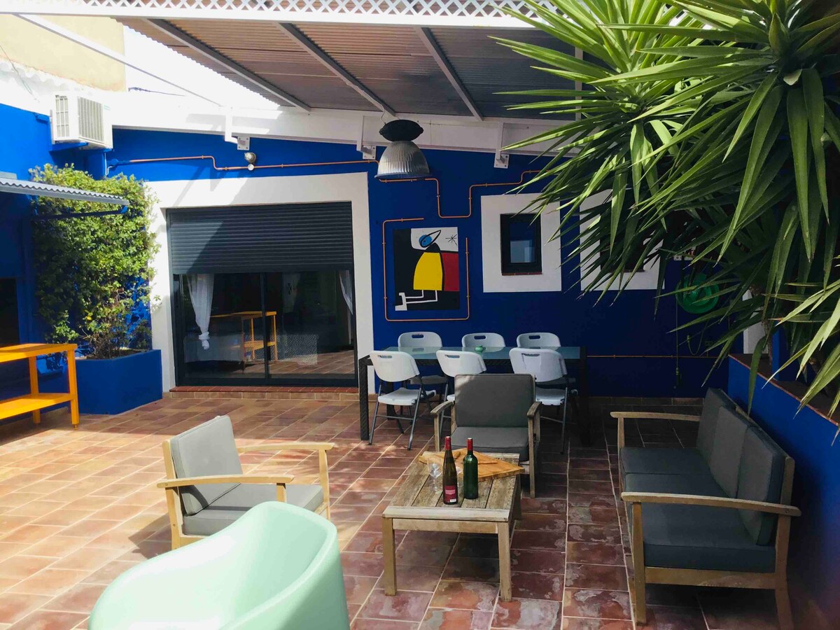 A vibrant outdoor space features a tiled patio with a mixture of modern and rustic seating options. A wooden table is surrounded by chairs, while lush greenery adds a natural touch. Colorful wall art and a shaded area enhance the inviting atmosphere.