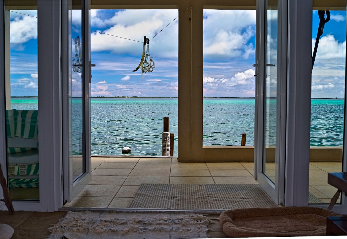 Double doors open to reveal a stunning view of calm waters, with vibrant turquoise hues stretching to the horizon. The floor is tiled and adorned with a textured rug. Sunlight highlights the serene atmosphere, inviting relaxation and connection with the surrounding nature.