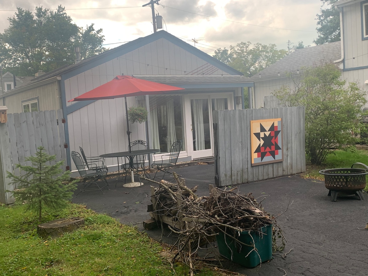 An outdoor space is presented with a patio featuring a round table and four chairs under a red umbrella. A fire pit is positioned nearby, with a pile of branches placed in the foreground. The area is enclosed by a wooden fence with decorative artwork visible.