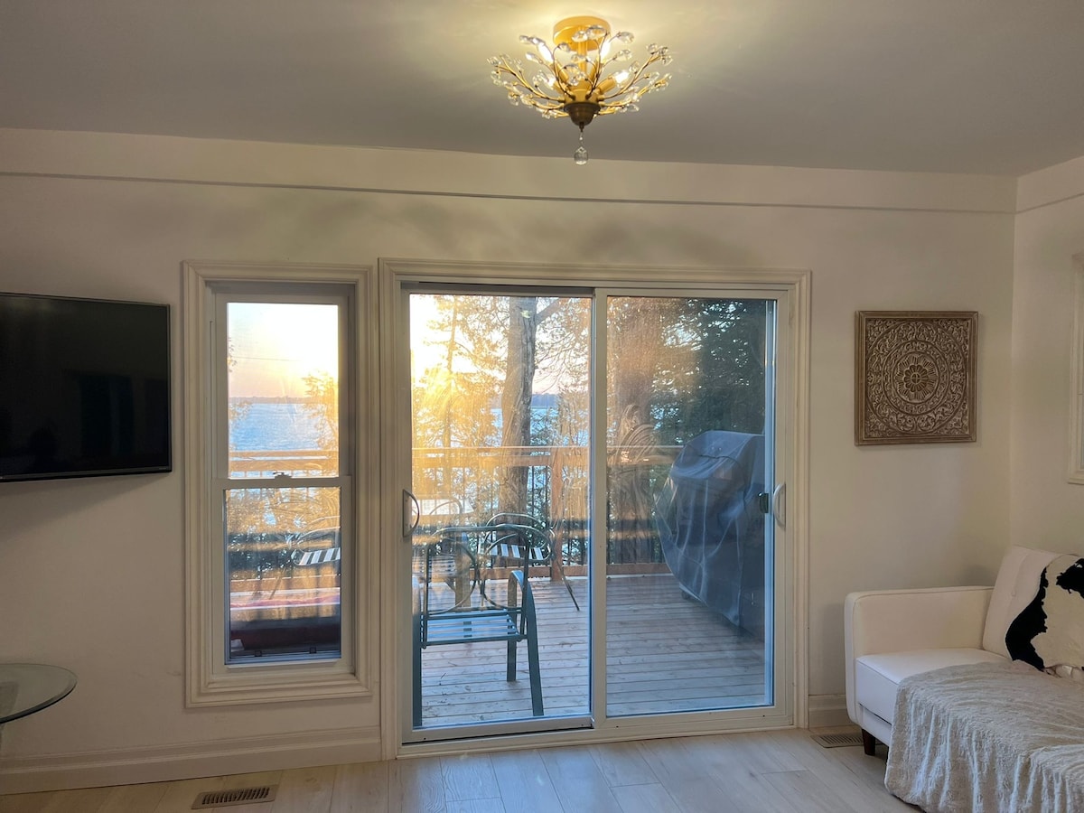 A bright living area features large sliding glass doors opening to a spacious deck, showcasing views of the bay. Natural light fills the room, illuminating light-colored walls and modern furnishings. A chandelier adds a touch of elegance, while a cozy sofa invites relaxation.