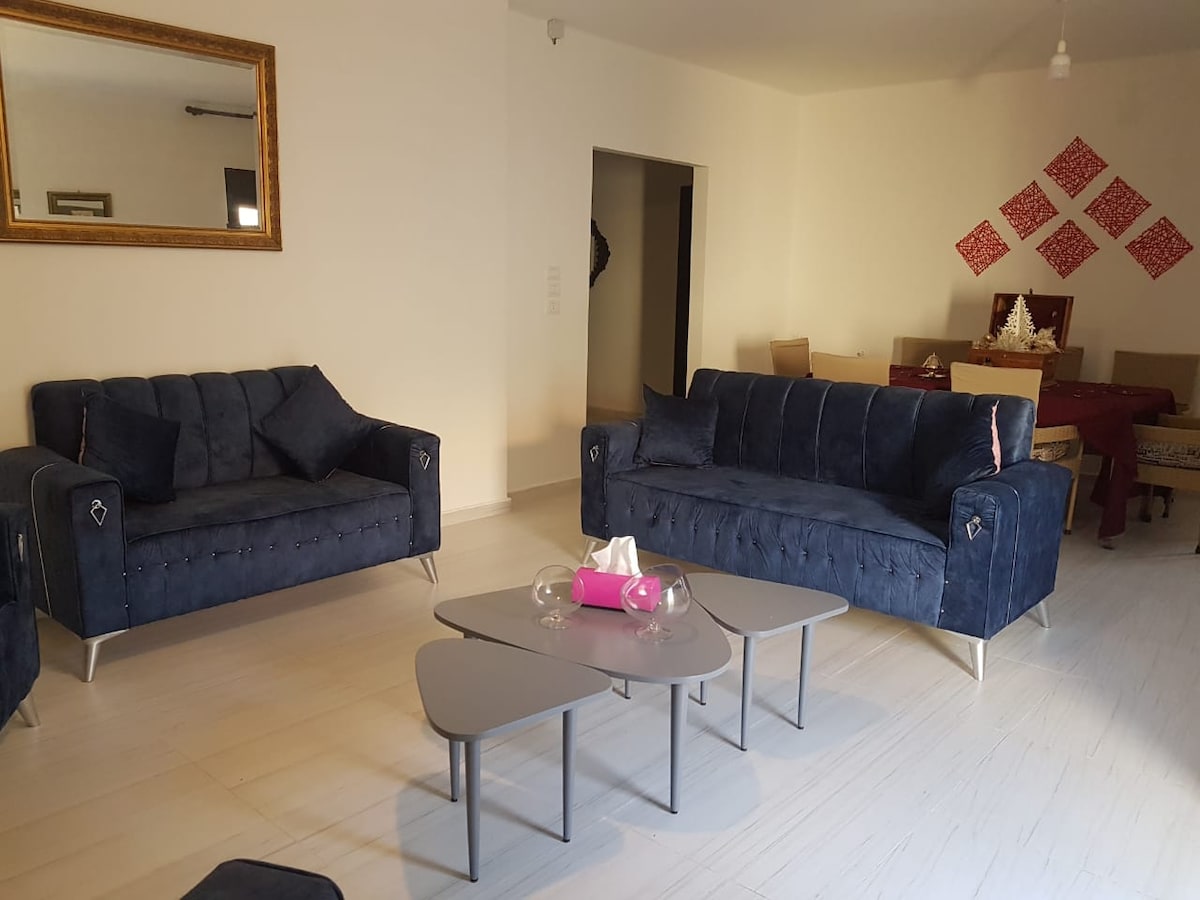 A spacious living area features two plush blue sofas and a modern coffee table. Light-colored walls enhance the brightness of the room, while a large mirror reflects the comfortable seating. A dining area with a table and chairs is visible in the background.
