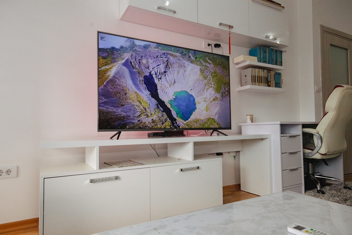 A modern living area features a large television mounted on a sleek white unit. Below the television, a set of storage drawers in a matching design provides ample space. A glass coffee table sits in front, and a comfortable chair is positioned nearby.