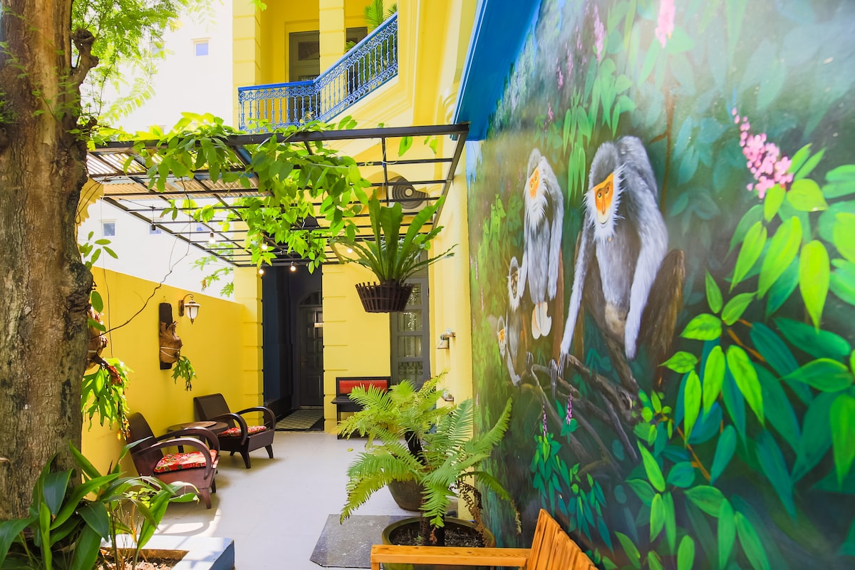 A vibrant outdoor area features a colorful mural depicting monkeys among lush greenery. The space includes wooden seating and hanging plants, creating a relaxed environment. The walls are painted in bright yellow, enhancing the cheerful atmosphere of this inviting garden area.
