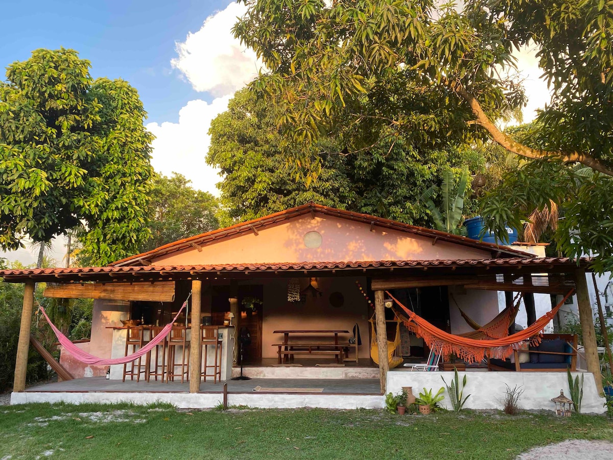 The home's exterior features a shaded porch with colorful hammocks and a spacious seating area. Natural light enhances the welcoming atmosphere, while lush greenery surrounds the property, providing a connection to nature. A rustic charm is evident in the wooden beams and open design.