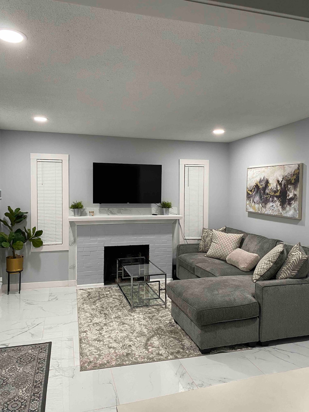 A cozy living area features a sectional couch arranged around a glass coffee table. The fireplace serves as a focal point beneath a wall-mounted television. Natural light filters through the shutters, and a potted plant adds a touch of greenery. Decorative artwork is displayed on the wall.