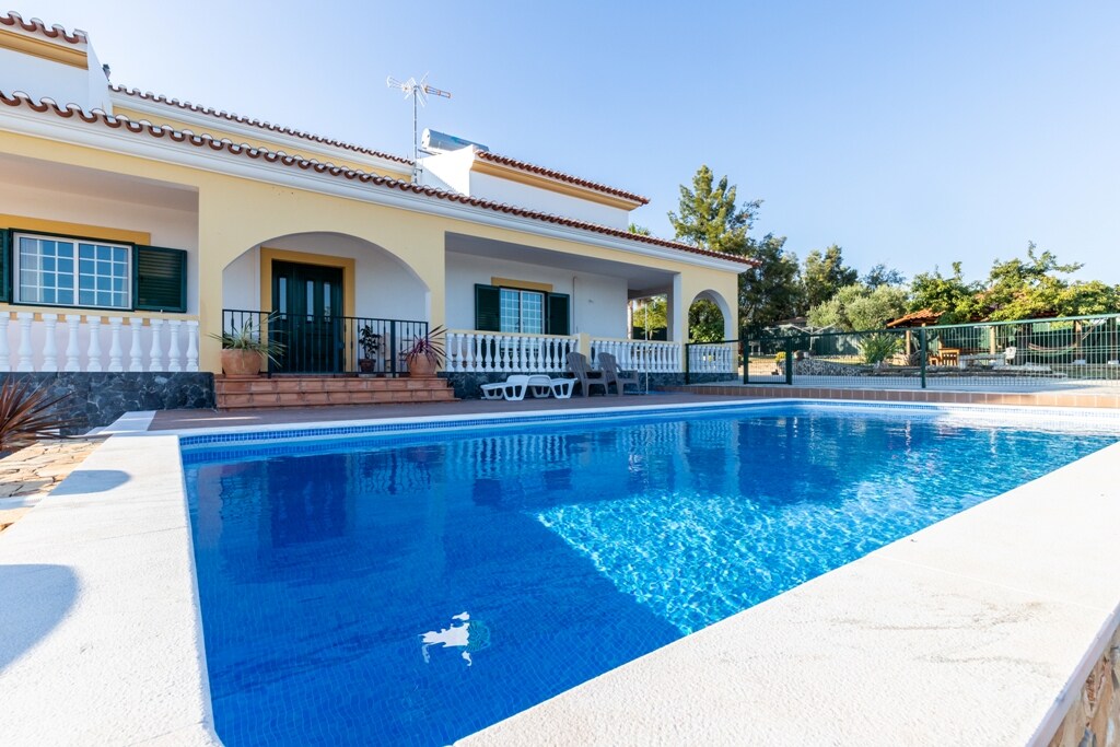 A clear blue salt-water pool is framed by a spacious terrace and surrounded by the home’s exterior, featuring a balcony with white railings. Sun loungers are positioned nearby, and lush greenery can be seen in the background, enhancing the outdoor ambiance.