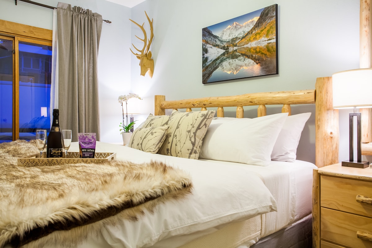 A comfortably arranged sleeping area features a spacious bed adorned with soft linens and decorative pillows. Natural light enters through a nearby window, highlighting a serene mountain landscape photograph above the headboard. A wooden deer antler accent adds a rustic touch to the decor.