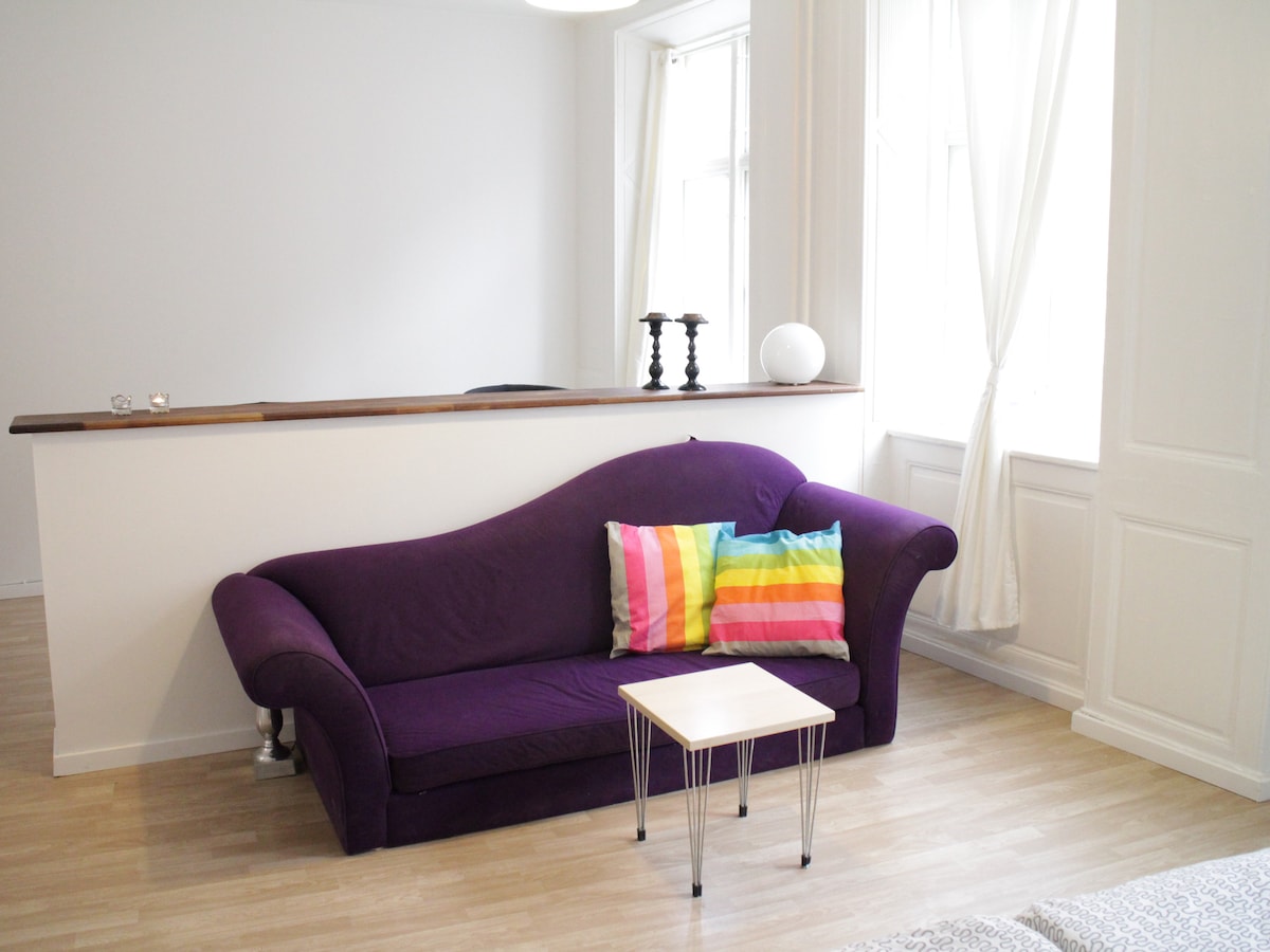A modern studio apartment features a plush purple sofa adorned with colorful striped cushions. A small white table sits in front of the sofa, while large windows allow natural light to illuminate the space. Light-colored wooden flooring complements the bright, airy atmosphere.