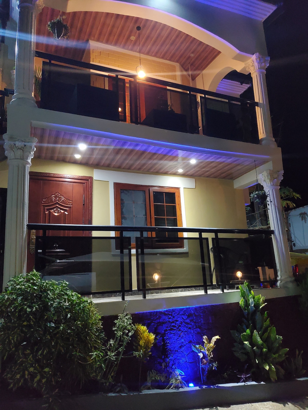 A two-story exterior view is presented, showcasing a beautifully lit balcony area. The entrance is highlighted by a decorative door, with two windows flanking it. Soft garden lights illuminate nearby greenery, adding a serene touch to the nighttime setting.