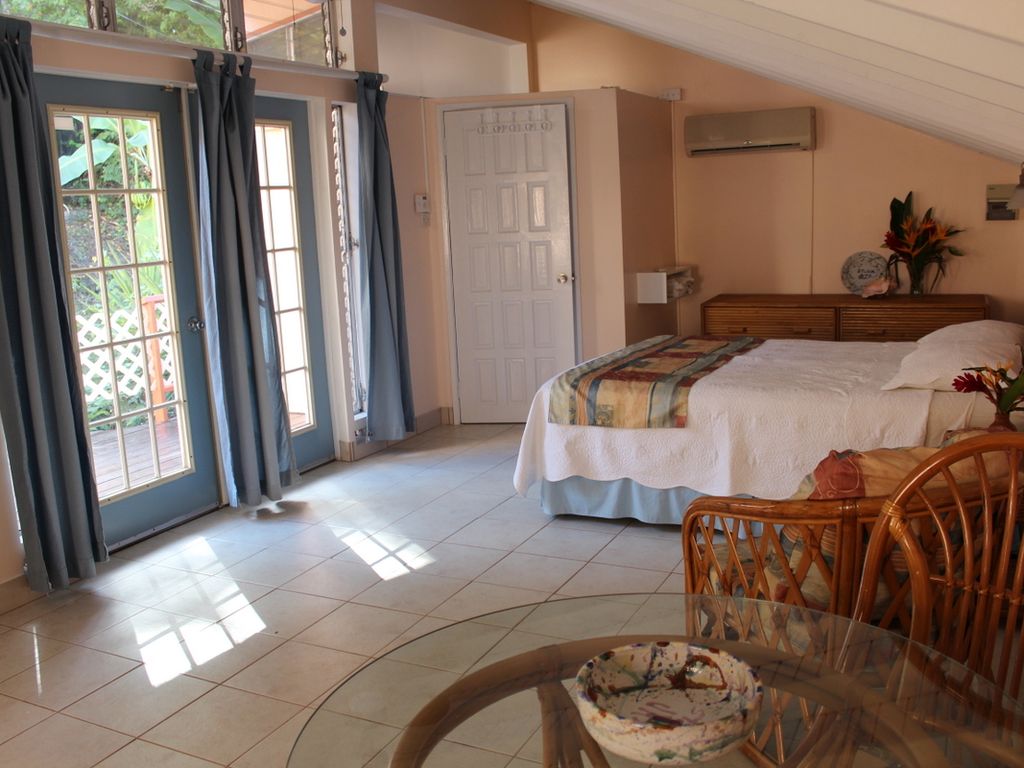 A spacious bedroom is highlighted by large, light-filled French doors leading to a private deck. Soft natural tones in the decor complement the wooden furniture, including a neatly made bed and a wicker dining set. High ceilings contribute to an airy atmosphere.
