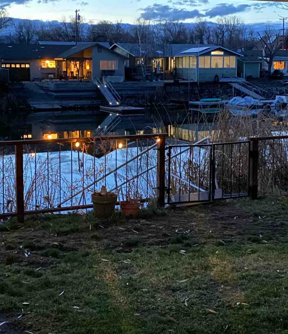 A view of the serene lake at dusk, with reflections of nearby houses visible on the water's surface. Soft lighting from the homes creates a warm ambiance. Tall grasses frame the scene, while pots are placed on the grass near the water's edge.