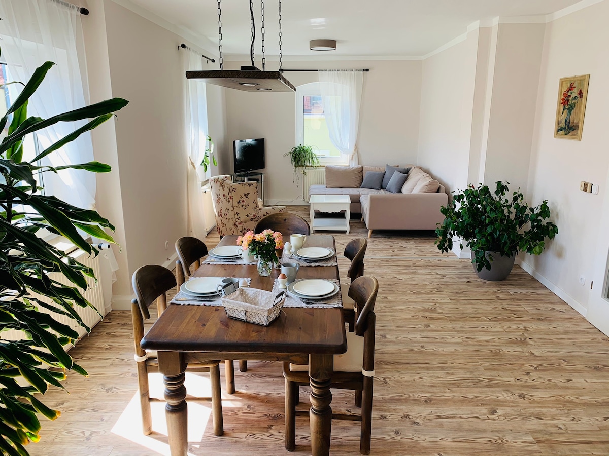 The spacious living area features a wooden dining table set for six, complemented by comfortable chairs. A cozy seating area with a light-colored sofa and a patterned armchair is visible. Natural light enters through large windows, enhancing the inviting atmosphere.