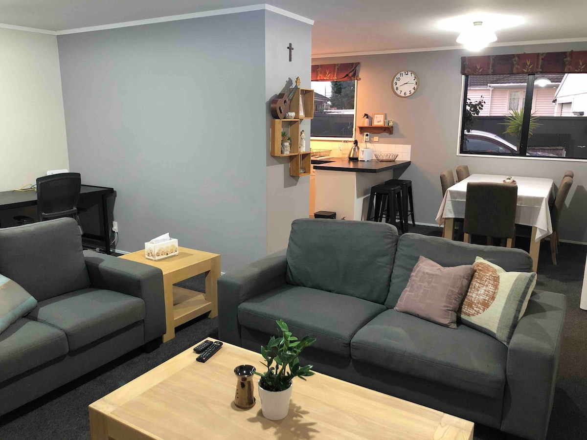 A comfortable living area features two sofas arranged around a wooden coffee table. Natural light enters through large windows, illuminating the space. In the background, a dining table and kitchen are visible, providing a functional gathering area.