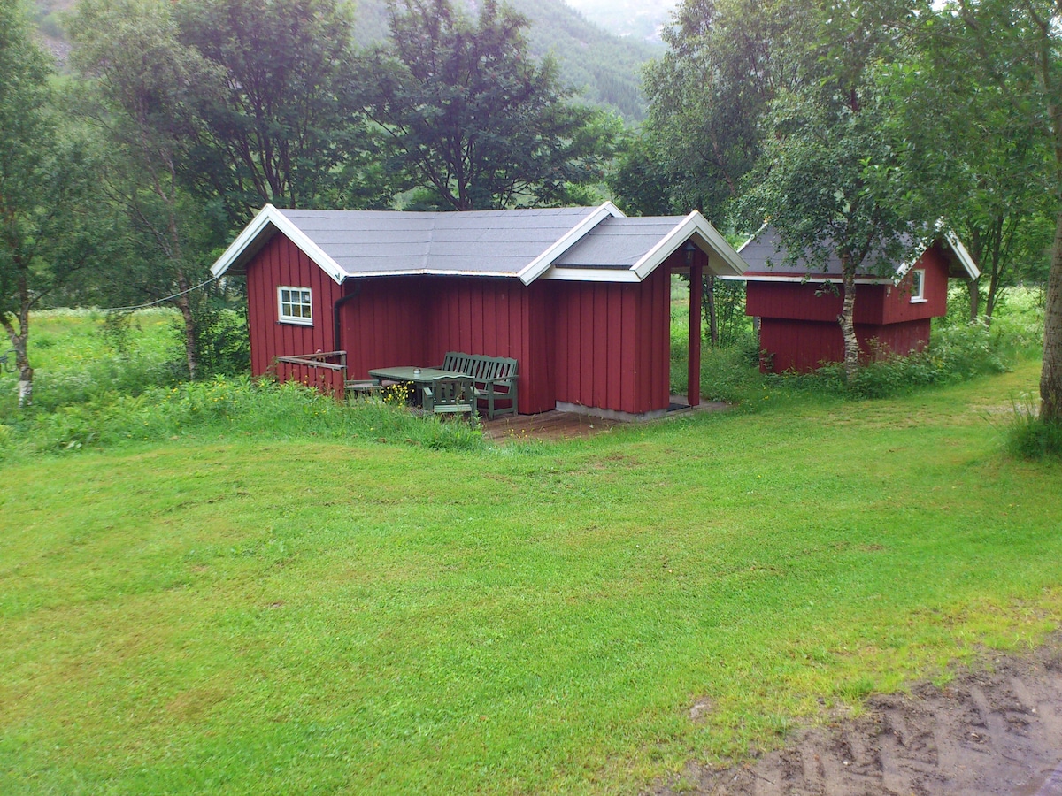 A small red cabin is set in a green lawn, surrounded by trees and hills. Outdoor seating is provided with a wooden table and benches. Another smaller cabin is visible in the background, blending harmoniously with the natural surroundings.
