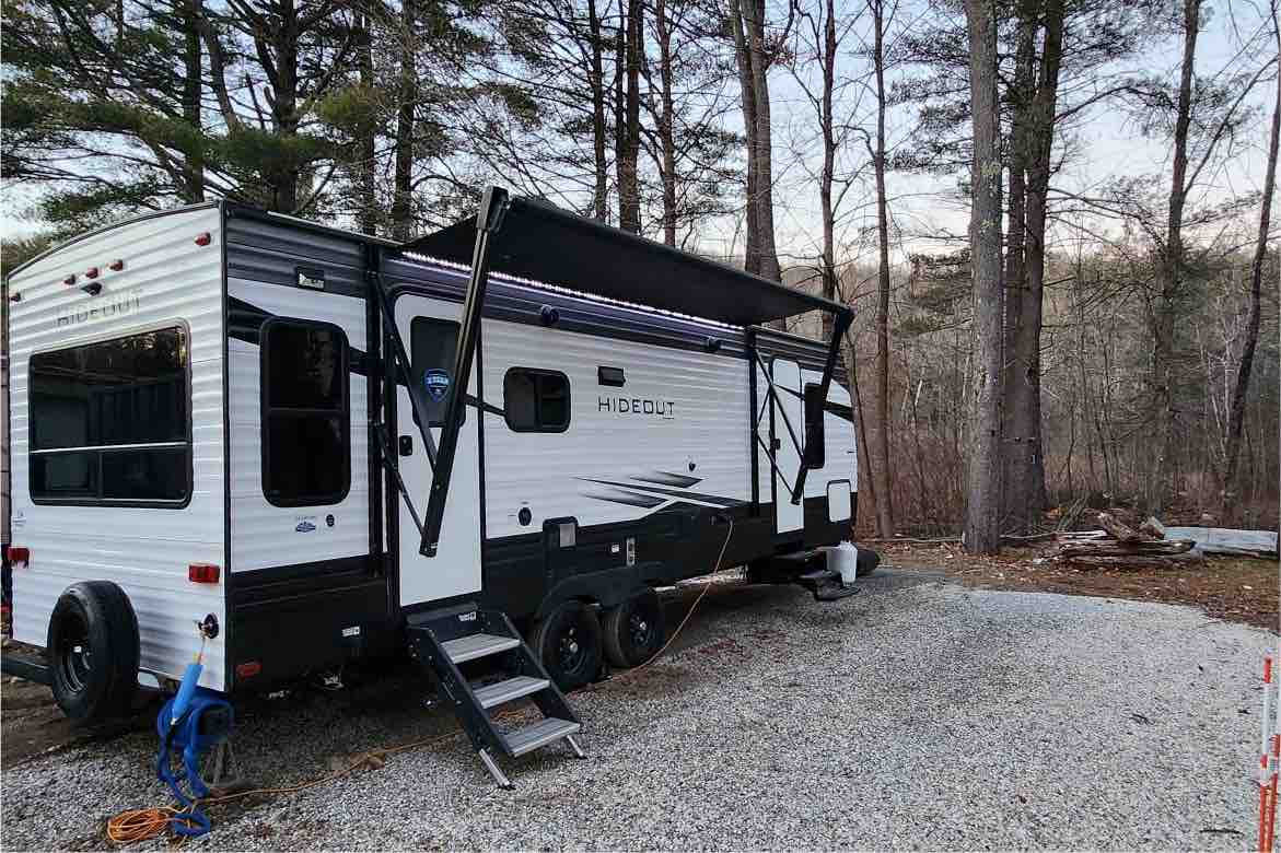 A modern travel trailer is positioned on a gravel area surrounded by tall trees. It features a retractable awning extending from the side, with a set of steps leading to the entrance. The exterior displays a sleek two-tone design in black and white.