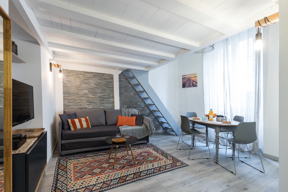 The living area features a comfortable sofa with decorative cushions and a stylish gray accent wall. A wooden table with modern chairs invites for dining. Natural light is provided by a large window draped with sheer curtains, complementing the warm tones of the rug underneath.