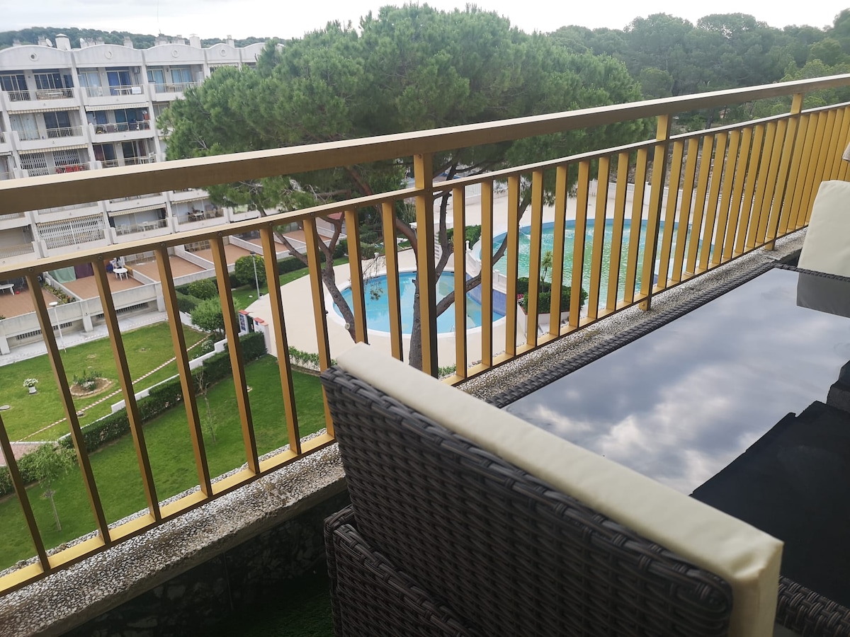 A balcony view overlooks a communal area featuring two swimming pools, grassy spaces, and trees. Sun loungers and a table with cushioned chairs are visible on the balcony, inviting relaxation while enjoying the surroundings.