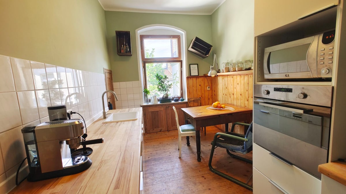 The kitchen features wooden countertops and a mix of light and dark cabinetry. A wooden dining table is accompanied by a green chair. A window allows natural light to fill the space, while modern appliances including a coffee maker and microwave are visible.