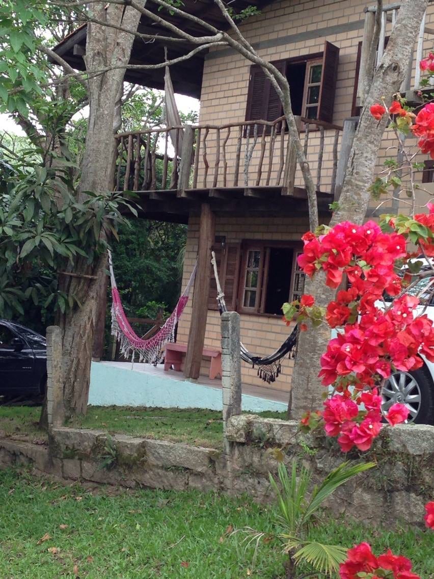 A two-story house is framed by lush greenery and flowering bougainvillea. Two hammocks are suspended from wooden beams on the balcony, inviting relaxation. A sturdy stone wall separates the property from a grassy area, while a parked car is visible nearby.