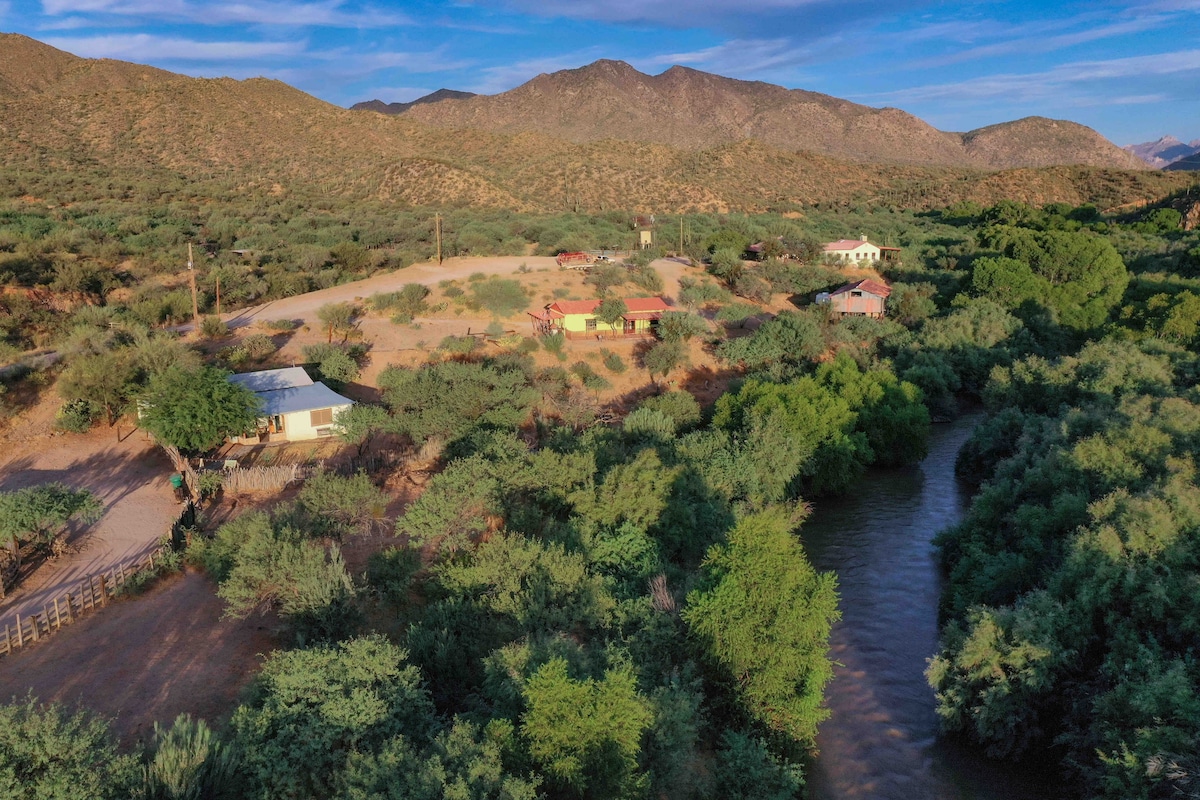 An expansive view captures a working cattle ranch surrounded by lush greenery and the Gila River, with rugged mountains in the background. Several adobe homes are visible amidst the landscape, promoting a sense of connection to nature and outdoor adventure.