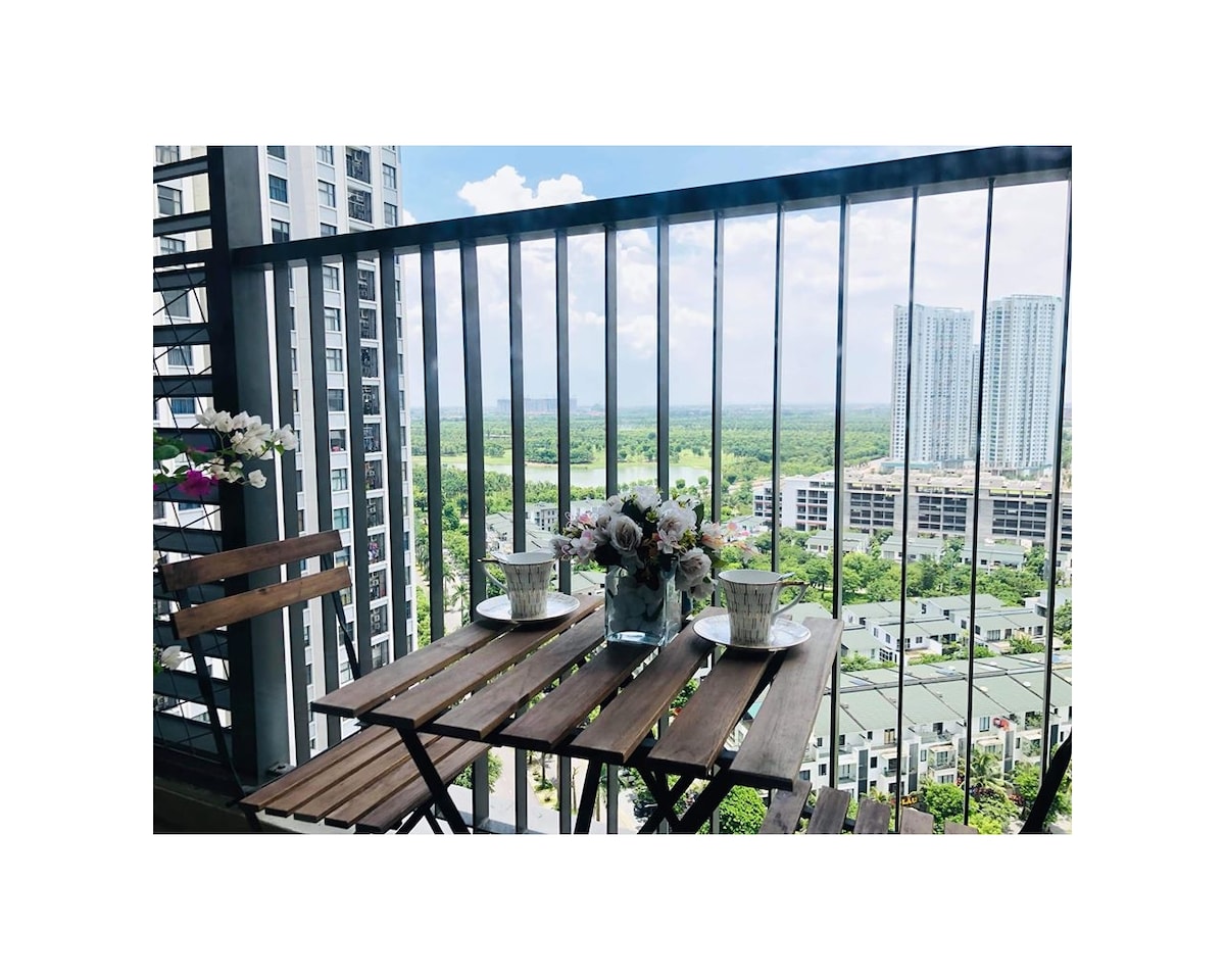 A small balcony is visible with wooden folding chairs and a matching table. Fresh flowers are arranged in a vase, providing a touch of nature. The view features a sprawling green landscape and tall buildings in the distance, under a bright blue sky.