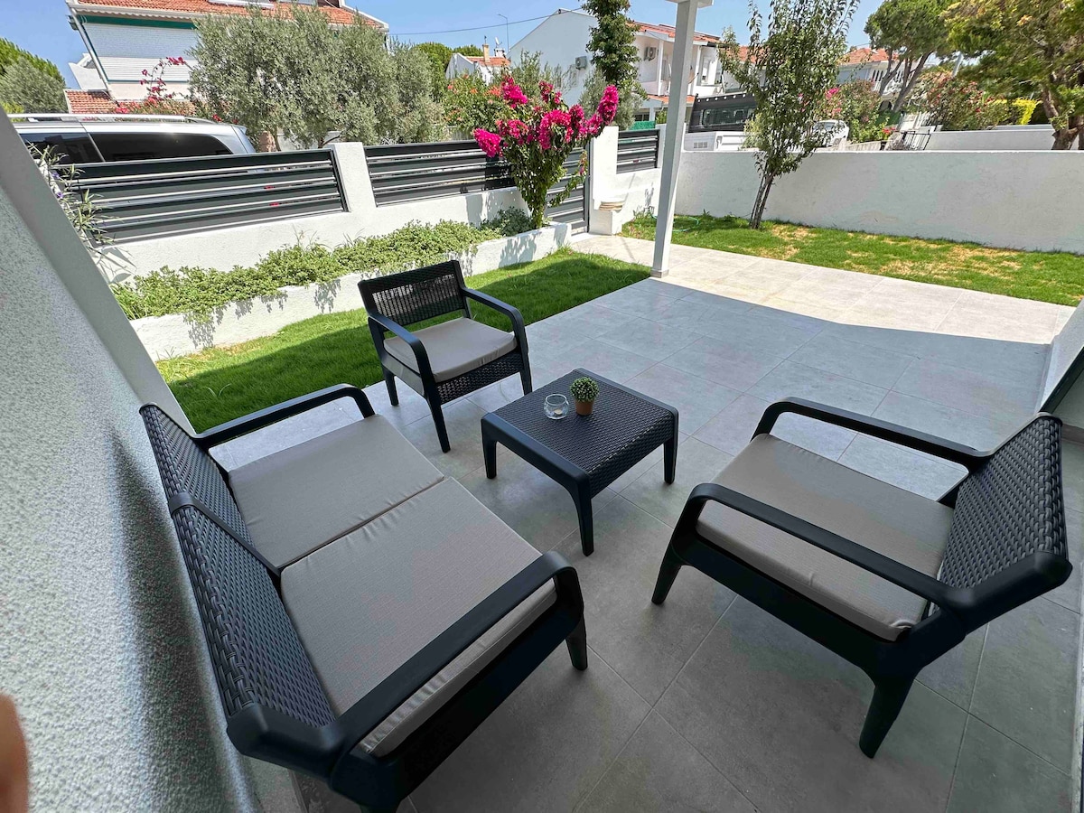 A covered outdoor seating area is furnished with two armchairs and a coffee table, surrounded by a well-maintained garden. Green grass and flowering plants add natural elements, while a paved pathway leads to the entryway, creating a serene space for relaxation.