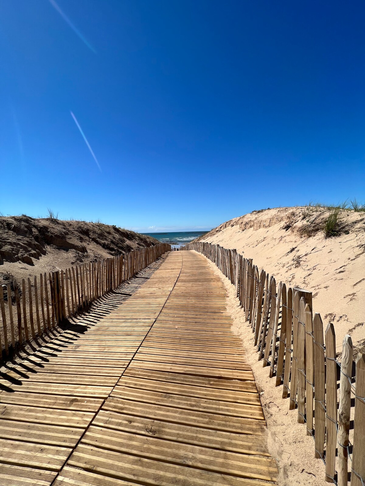 A wooden pathway leads towards the beach, flanked by sandy dunes on either side. Clear blue skies are visible above, enhancing the serene coastal environment. The sound of waves can be imagined in the distance, creating an inviting atmosphere for beachgoers.