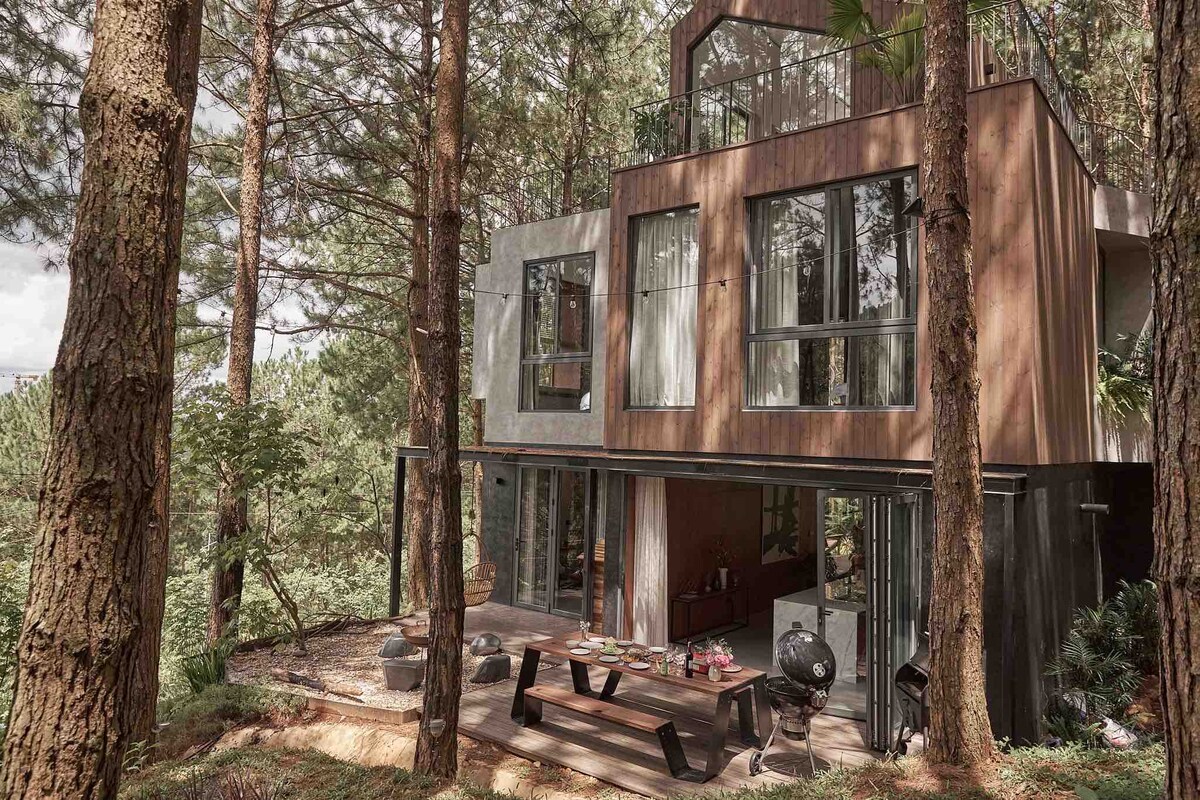 A modern full house is nestled among tall pine trees. Large glass windows invite natural light into a spacious outdoor deck, featuring a dining area and a BBQ pit. The blend of wood and concrete materials emphasizes the home's contemporary design.
