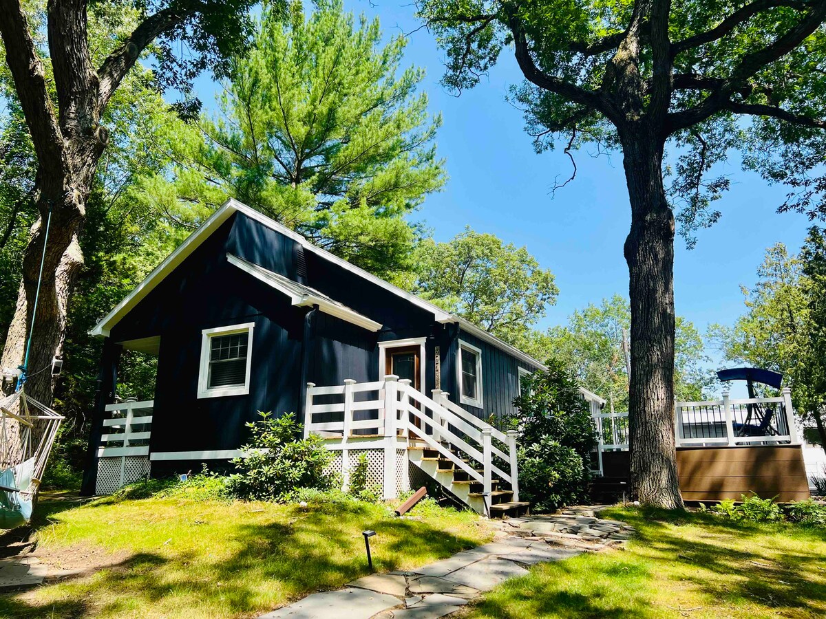 A cozy cottage with dark blue siding is framed by tall trees and lush greenery. A white railing leads to the entrance, and a wooden deck with outdoor seating is visible at the side. A stone pathway gently winds through the grassy yard.