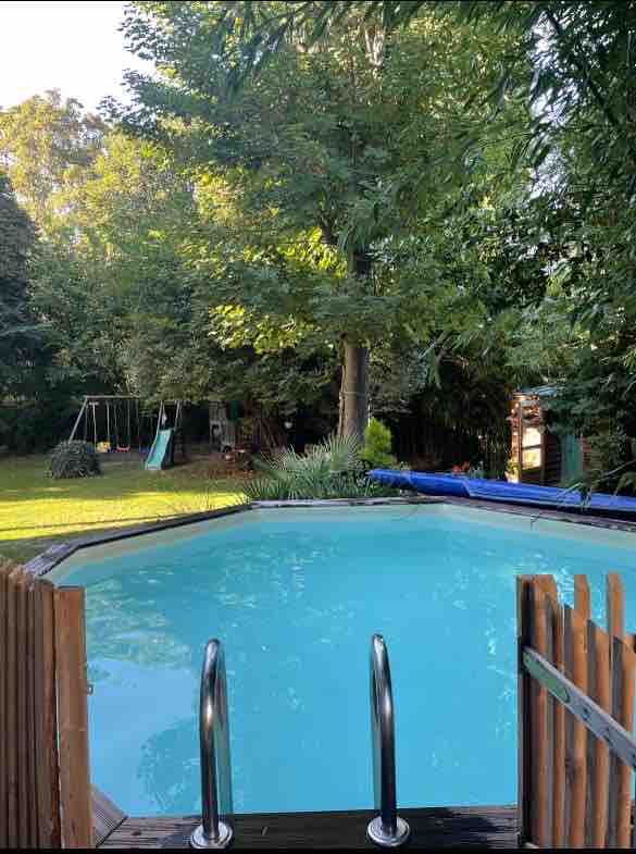 A private pool with a wooden deck is surrounded by lush greenery and trees. The vibrant blue water is inviting, while a grassy area includes a swing set and a slide, creating a playful outdoor space.