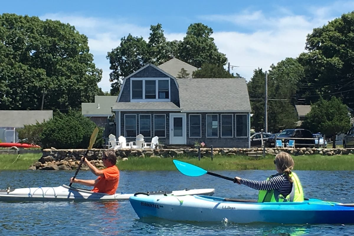 A view of a rustic cottage is presented against a backdrop of greenery, featuring white chairs on a deck. Two individuals are seen kayaking in the foreground, with clear blue water reflecting the building's muted grey tones. The sky is bright, adding to the outdoor atmosphere.