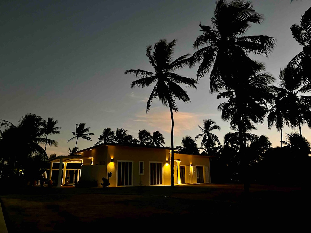 The villa is illuminated under a twilight sky, surrounded by numerous palm trees. The structure showcases large glass doors and windows, reflecting the soft glow of outdoor lighting. The tranquil atmosphere invites relaxation, set amidst a serene natural landscape.