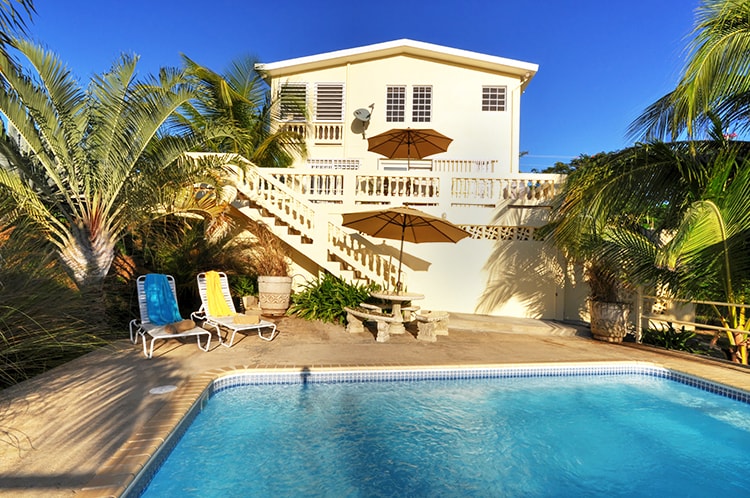 A spacious outdoor area features a private pool surrounded by tropical landscaping. Two lounge chairs in yellow and blue sit beside the pool, while a shaded table and chairs provide a relaxing space. The two-story house stands in the background, with clear skies above.