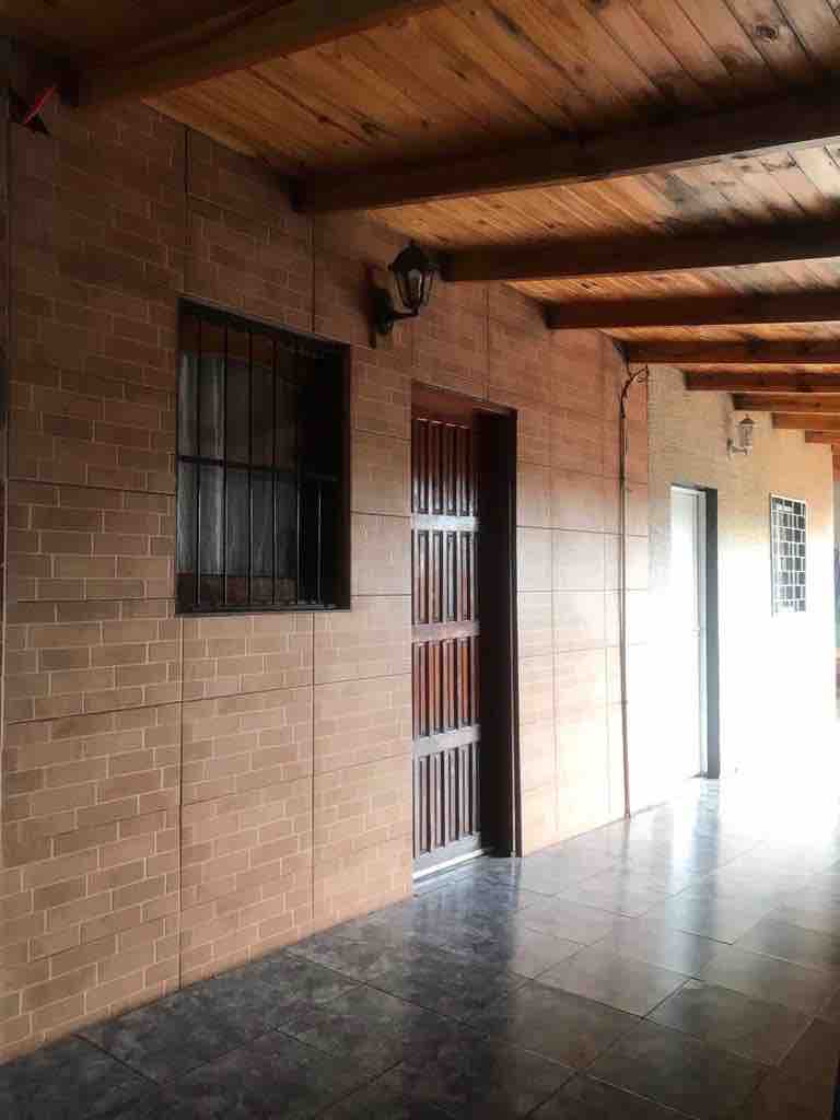 A corridor features a tiled floor and wooden ceiling beams. Solid wooden doors are framed by brick walls, with window grilles providing security. Natural light enters the corridor from the end, creating a bright pathway to the rooms.